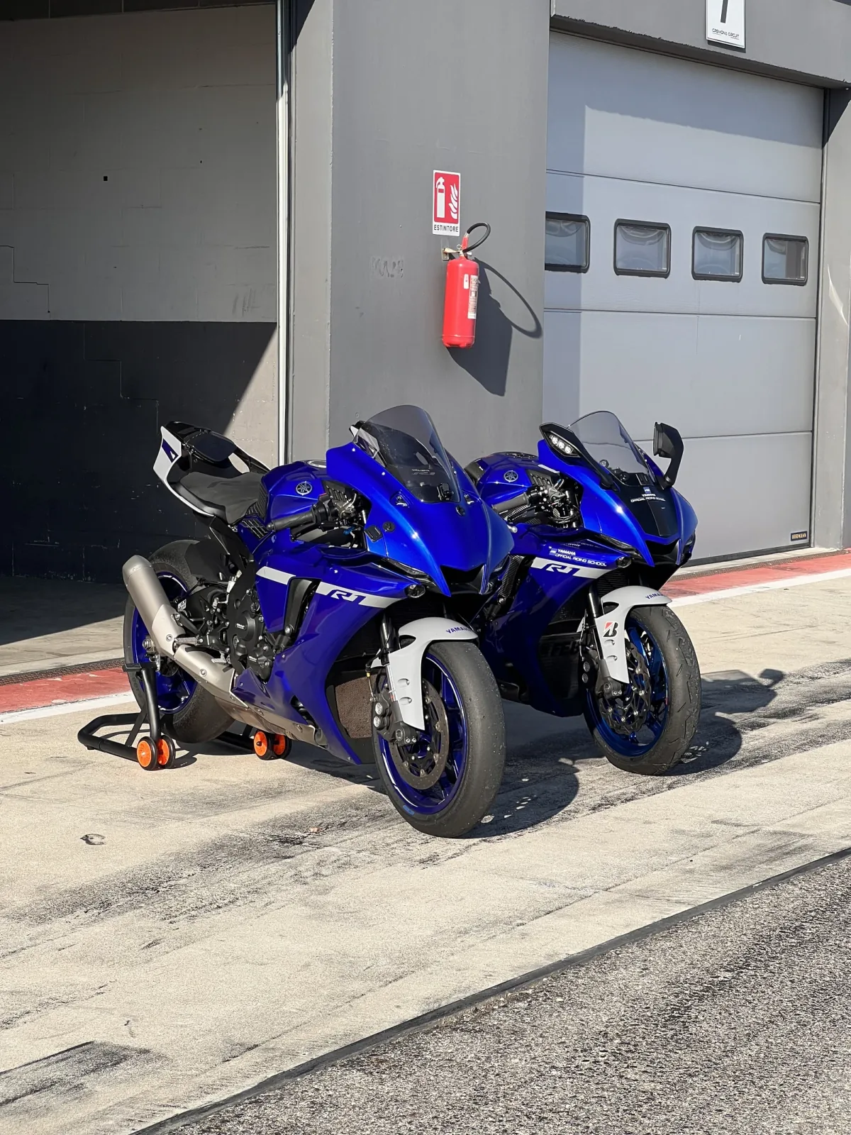 Two Yamaha R1 on paddock stands in front of a garage in the Cremona Circuit pit lane.